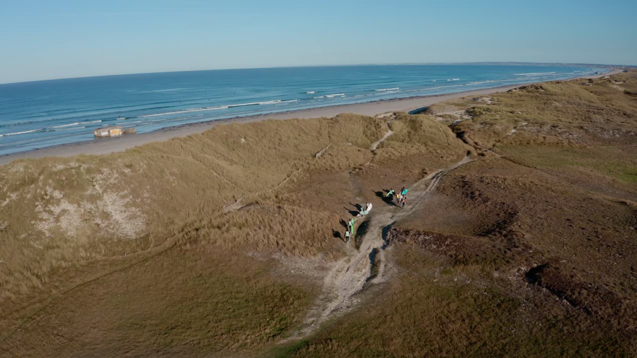 vista de avión no tripulado de surfistas caminando a través de dunas de arena hacia el océano bajo un cielo azul claro