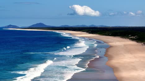 Una-Playa-Con-Una-Orilla-Arenosa-Y-Una-Playa-Arenosa-Al-Lado-Y-Un-Océano-Azul-Con-Olas-Blancas-A-Vista-De-Pájaro