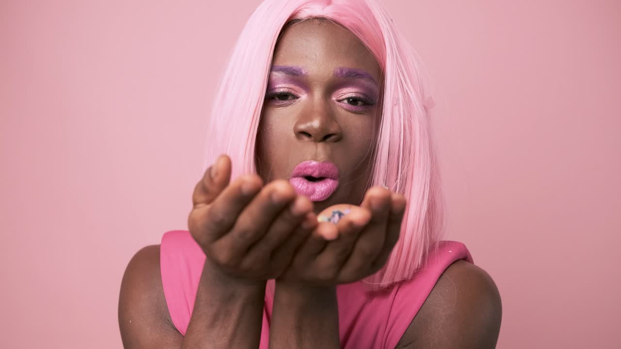 Transgender african person with pink dress blowing confetti on pink background