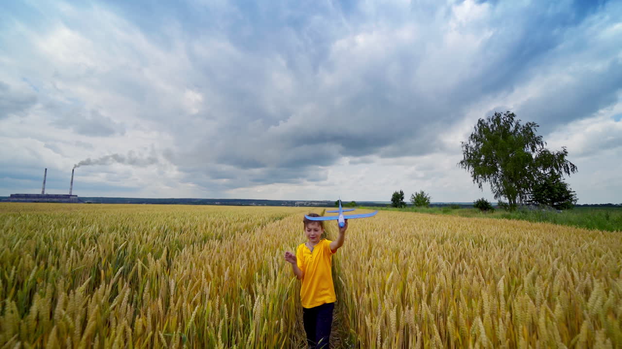 Child plays outdoors. Cute boy walks on yellow field and imitates flight with his toy plane under clouded sky in summer.
