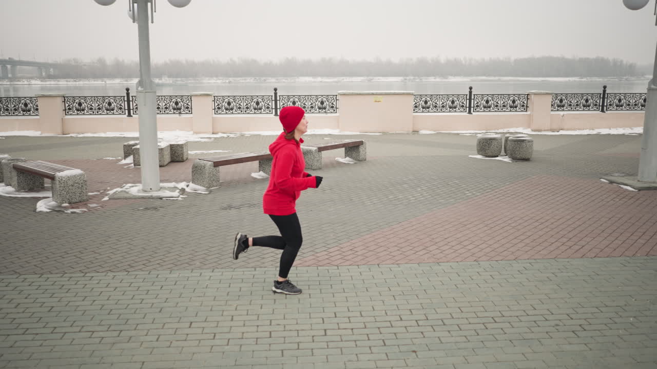 Side view of woman jogging outdoors during winter on interlocked pavement, scenic urban environment with benches, decorative railings, snow-covered ground, and river setting under cloudy skies