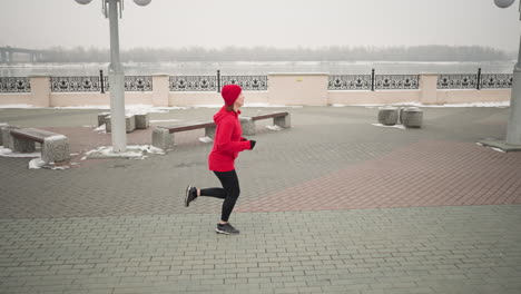 side view of woman jogging outdoors during winter on interlocked pavement, scenic urban environment with benches, decorative railings, snow-covered ground, and river setting under cloudy skies