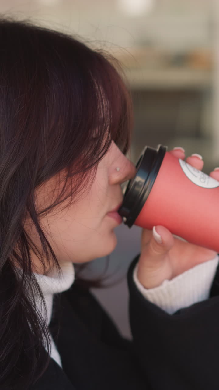 검은 코트를 입은 여성이 빨간 컵에서 커피를 마시고, 만족스러운 미소를 지으며 부드럽게 내려 놓고, 흐릿한 도시 배경으로 편안하고 도시적인 느낌을 향상시니다.