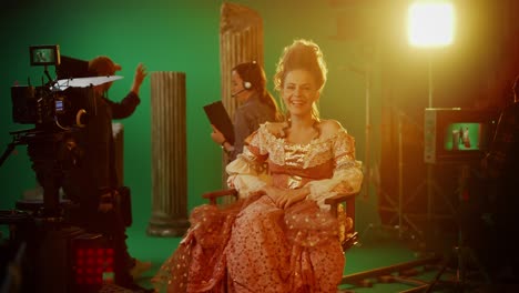 on period costume drama film set: beautiful smiling actress wearing renaissance dress, sitting on a chair happily with green screen in the background. studio shooting history blockbuster