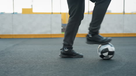 close-up leg view of athlete skillfully training with football in sport arena, showcasing control and precision while tapping ball forward, background features chain-like fence and entrance