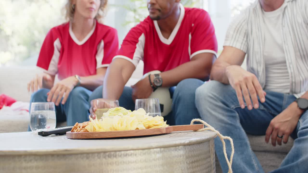 Tifosi diversi con maglie rosse seduti sul divano che guardano una partita in TV, allungando la mano per prendere le patatine, con un pallone da calcio
