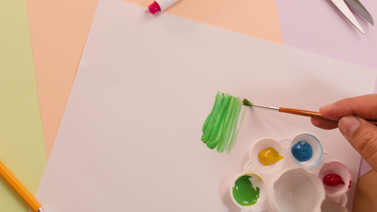 Artist hand paints green strokes on white paper using brush, palette, and primary colors overhead