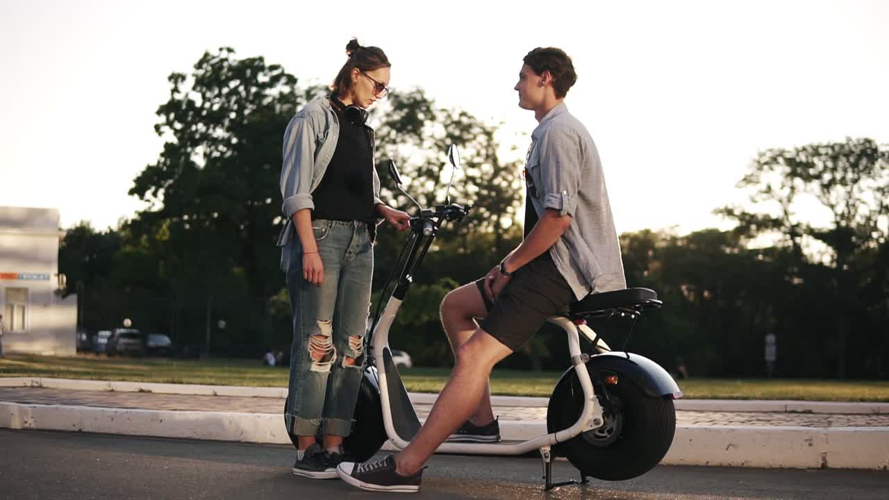Young male and female are have time together outdoors. Boy's sitting on an electro mini bike and smiling. The girl in sunglasses stands near and talks. Sun shines on the background
