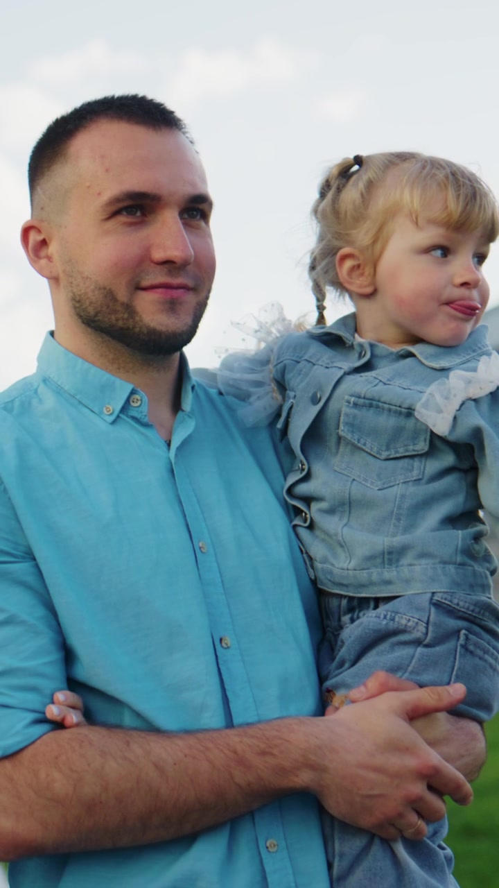 Un uomo tiene in braccio un bambino/a piccolo/a in un campo aperto. Il bambino/a indossa un completo di jeans con le trecce. L'uomo guarda dolcemente la fotocamera. Il bambino/a guarda altrove con un dolce sorriso. La scena cattura un tenero momento padre-figlia.