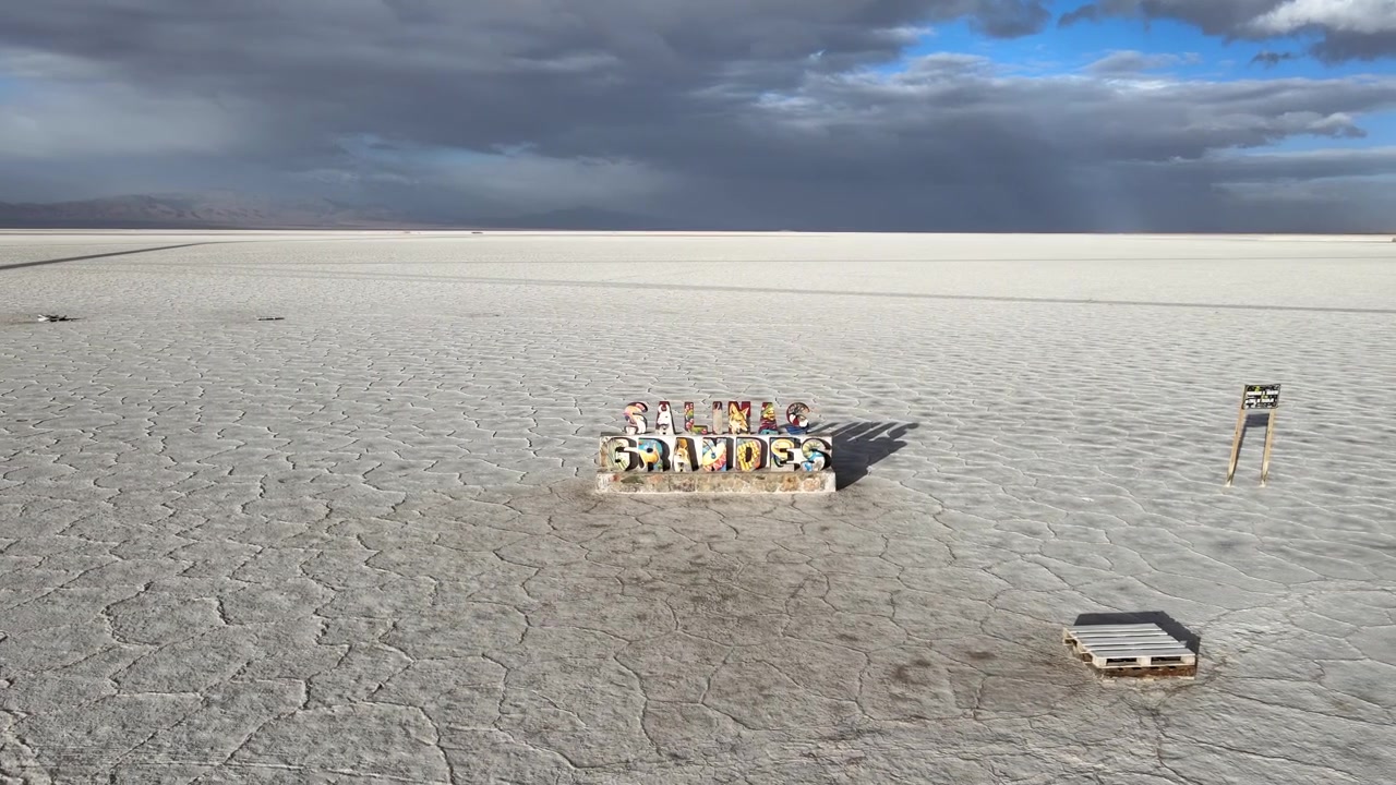 Colorful Salinas Grandes sign stands alone on cracked white salt flats under dramatic clouds in Jujuy, Argentina, seen from a moving drone.