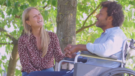 Una-Pareja-Madura-Sonriente-Con-Un-Hombre-Sentado-En-Silla-De-Ruedas-Hablando-Juntos-En-El-Parque