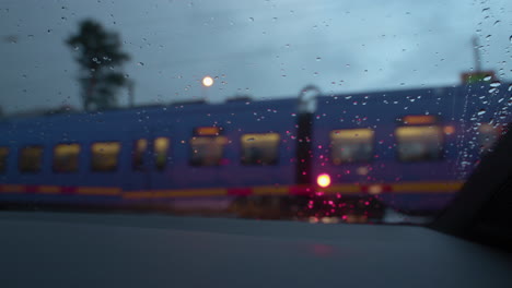 Close-up-detail-shot-of-the-rain-droplets-on-the-wind-shield-of-a-VW-Golf-car-as-a-purple-electric-train-commuter-drives-by-from-right-to-left-at-a-train-station-in-Europe-on-a-moody-dark-rainy-day