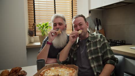retrato de una feliz pareja de gays de mediana edad un hombre con cabello gris y una barba exuberante junto con su novio moreno con paja en una camisa a cuadros come pizza durante su almuerzo en casa en la cocina