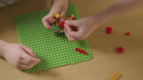 Overhead-Shot-Of-Two-Children-Playing-With-Plastic-Construction-Bricks-On-Wooden-Table-At-Home