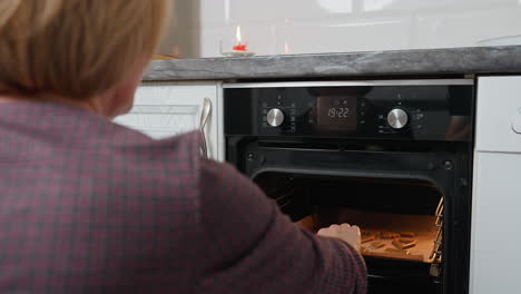 rear view of woman checking cookies in oven, closing oven door back after inspection, red candle burning on kitchen counter in modern kitchen with warm cozy vibe