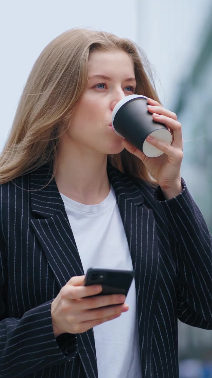 Ung forretningskvinde bruger smartphone og drikker kaffe udendørs.