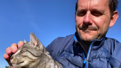 Primer-Plano-De-Un-Hombre-Adulto-Sonriente-Abrazando-A-Un-Gato-Afuera-Bajo-El-Sol-En-El-Cielo-Azul