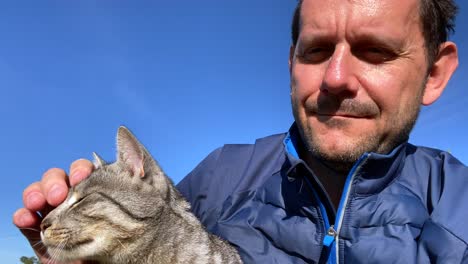 closeup shot of smiling adult man cuddling cat outside in the sunshine at blue sky