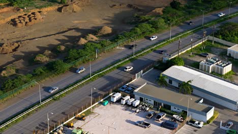 Paralaje-Aéreo-Alrededor-De-Automóviles-Que-Circulan-Por-Una-Autopista-Tropical-Junto-A-Edificios-De-Fábricas-Industriales