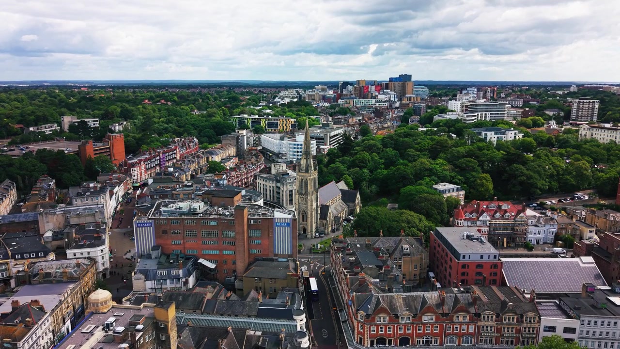 Luftfoto fra drone af Bournemouth centrum, der viser et fremtrædende kirketårn, historiske bygninger, moderne udviklinger, gader, grønne parker og den bredere bysilhuet under en overskyet himmel.