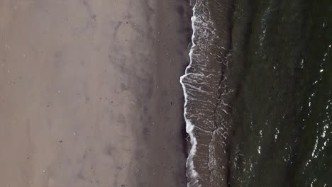 A-top-down-view-over-an-empty-beach-as-the-waves-gently-crash-onto-the-shore-in-slow-motion