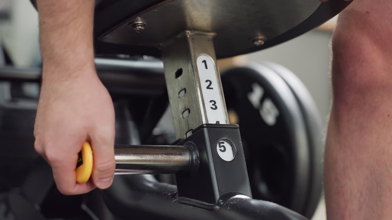 Man uses yellow pull pin on weight bench to adjust seat height, hand gripping metal lever as mechanical pointer moves to numbered position, showcasing gym equipment detail under studio lighting