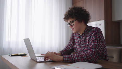 un hombre rizado con una mirada seria trabaja en una computadora portátil sentado en una cocina moderna. joven estudiante independiente usando computadora portátil estudiando en línea trabajando desde casa en internet