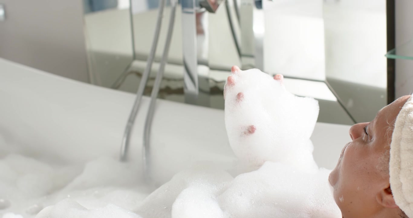 Una mano emerge dalla schiuma nella vasca da bagno, modellando le bolle in un'alta colonna vicino al rubinetto cromato.