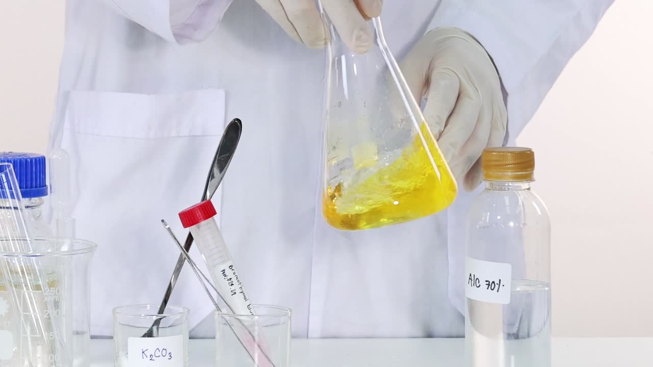 A scientist in gloves stirs a yellow liquid in a flask, surrounded by lab equipment.