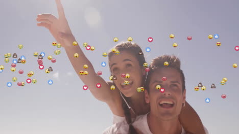 man giving piggyback ride and woman pointing upward on beach with floating social media emojis