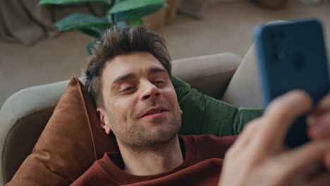 cheerful man texting smartphone lying on home couch closeup. smiling guy texting