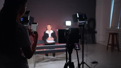 professional camera recording a lady sitting at desk and talking. unrecognized man fixing the camera display. long-haired girl takes video on her phone.