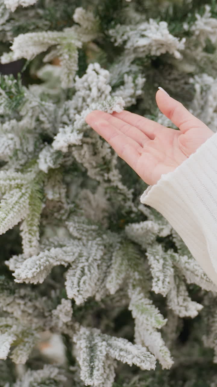 Close-up rear view of young lady in white sweater gently touching snow-like Christmas tree in decor shop, focus on hands interacting with frosty branches