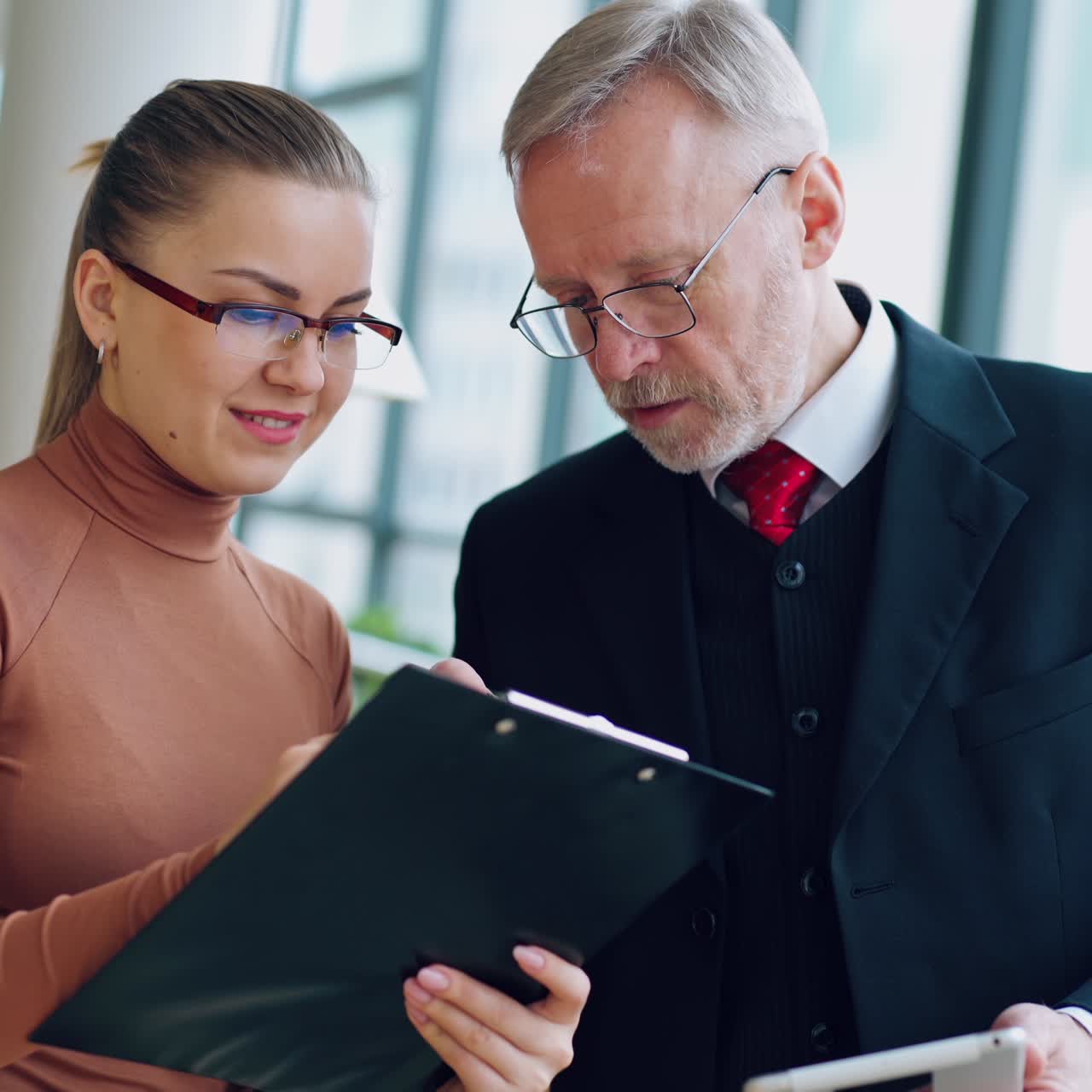 Young woman assistant and senior businessman working together in a light room with big windows, laughing. Team work. Business video