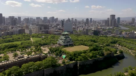Aerial-of-Osaka-Castle-with-park,-moat,-skyscrapers,-and-city-in-Osaka,-Japan