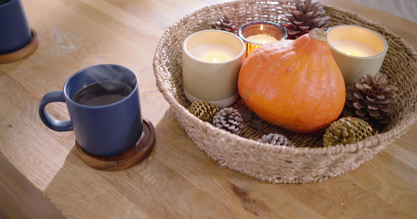 La cámara se desliza y hace zoom para resaltar una cesta tejida, velas encendidas y una taza humeante sobre una mesa de madera