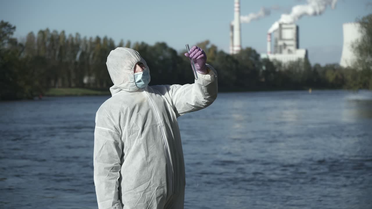 человек в защитном костюме держит пробирку с речным образцом, вид снаружи со среднего плана, промышленный фон
