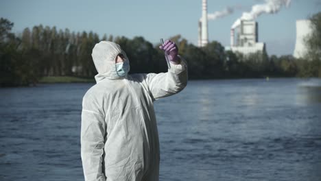 Person-wearing-protective-suit-holding-test-tube-with-river-sample,-outside-middle-shot-view,-industrial-background