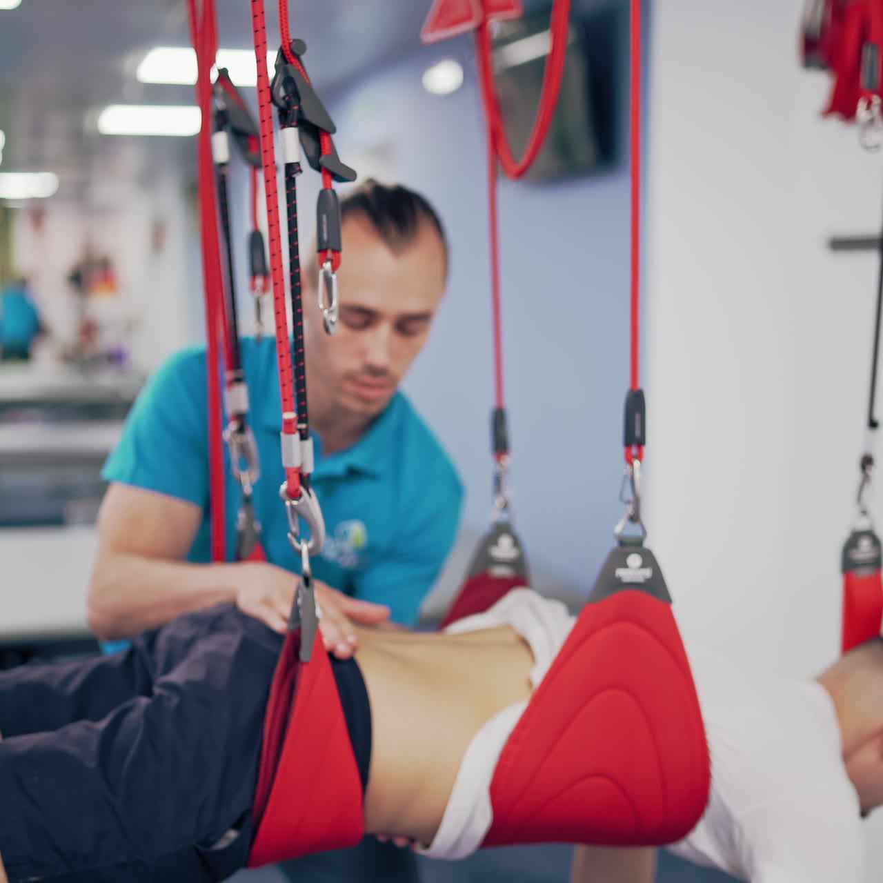 Client male is undergoing rehabilitative physiotherapy on a special suspension rope system. Young man is doing workout on a new equipment ropes with the help of the trainer. Modern rehabilitation center.