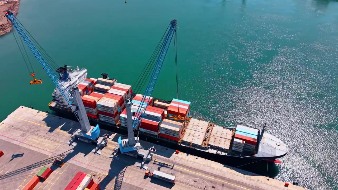 Burgas, Bulgaria, 1 May 2026: Container ship berthed with cranes and harbor water. Drone view of container ship at quay, two blue cranes, turquoise water, sun reflections and open copy space.