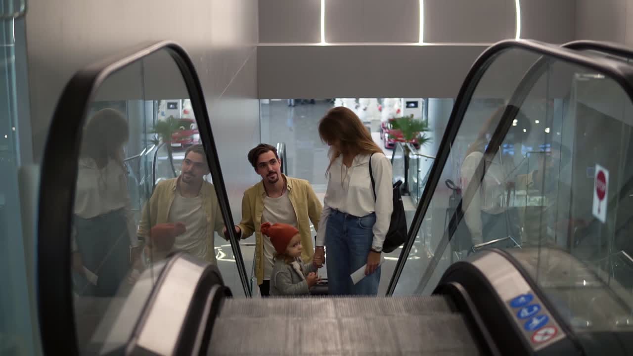 Young attractive caucasian family with suitcase going up by the escalator with their pretty daughter hold by mother's hand at the