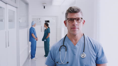 Mature-Male-Doctor-Wearing-Scrubs-Standing-In-Busy-Hospital-Corridor-With-Colleagues-In-Background