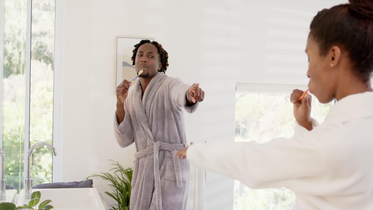 African American couple at bathroom sink in robes, man turning to woman, playfully brushing teeth