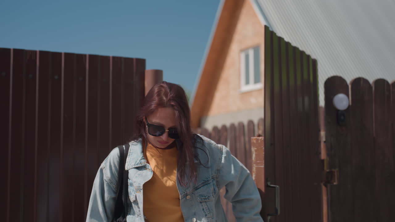 Young woman in red shirt opens metal gate while two friends follow closely behind on sunny day, entering private property with final person preparing to close gate