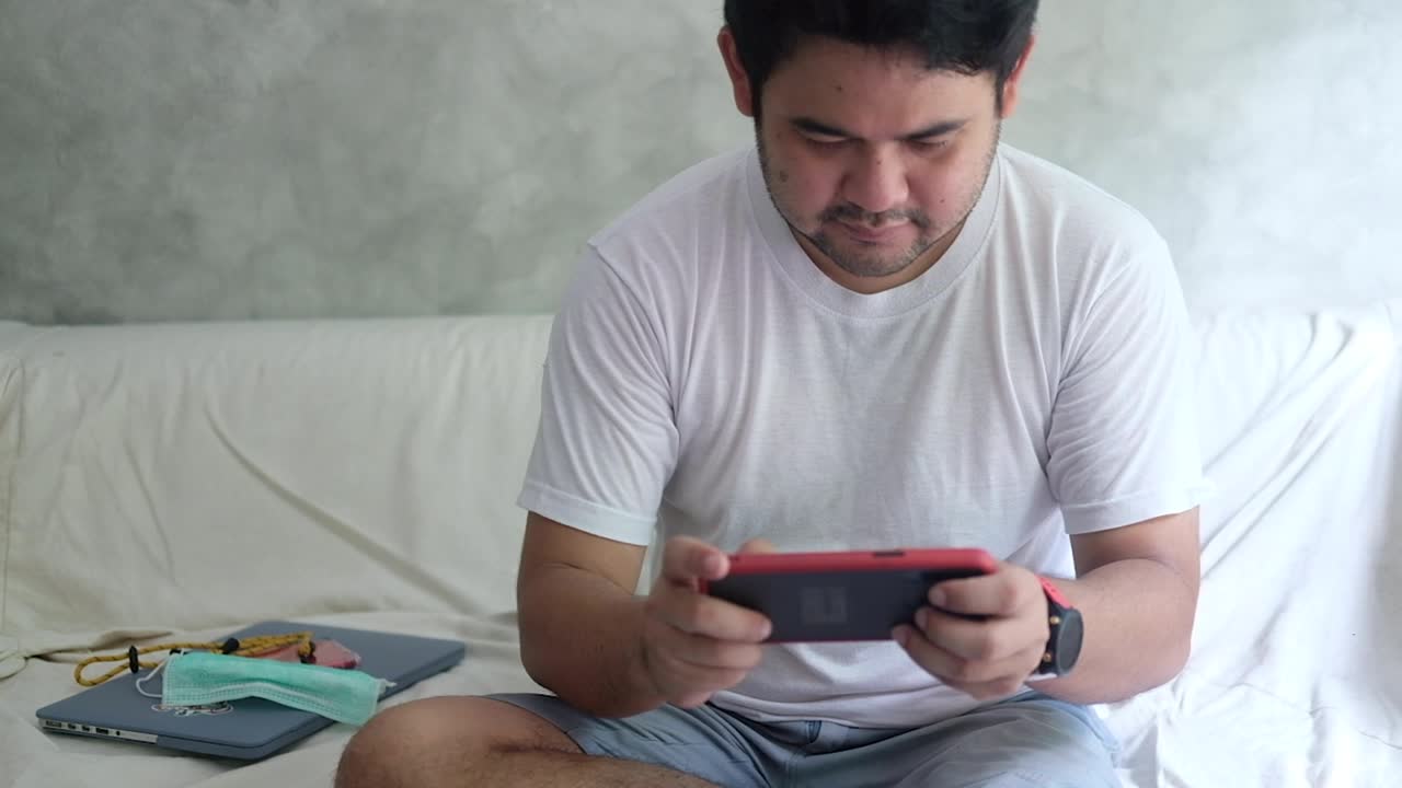 Young bearded man sit on the sofa playing mobile game on his smartphone, self quarantine concept