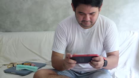young bearded man sit on the sofa playing mobile game on his smartphone, self quarantine concept