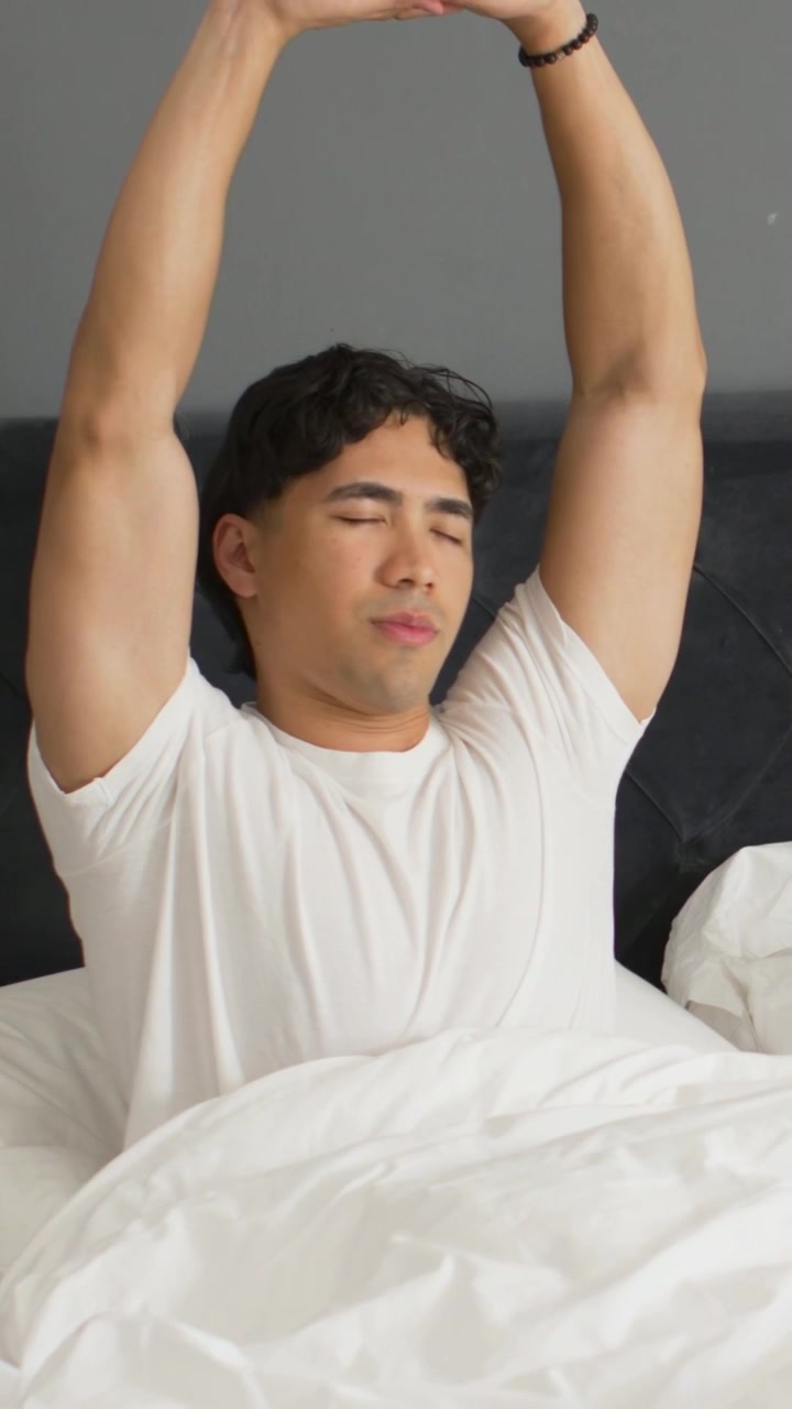 Video verticale: Uomo che si sveglia, seduto in camera da letto, che tira il piumone per lo stretching mattutino, con un braccialetto.