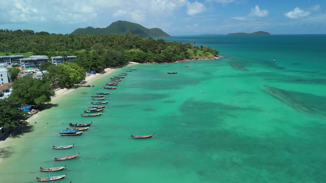 Drone aereo scende su Rawai Beach, mostrando barche longtail allineate lungo l'acqua turchese, edifici costieri, colline verdi e isole vicine a Phuket