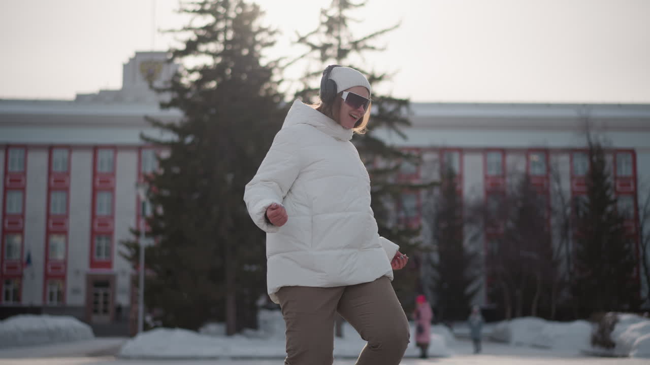 Teenage girl wearing white winter coat, beanie and headphones jumps and dances on paved snowy plaza, throwing hands side to side and up and down, grooving to music with excitement under cold daylight