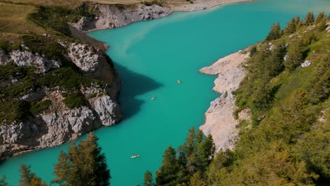 Aerial-view-of-kayakers-enjoying-the-serene-turquoise-waters-of-Mont-Cenis-Lake,-surrounded-by-lush-greenery-and-rocky-cliffs